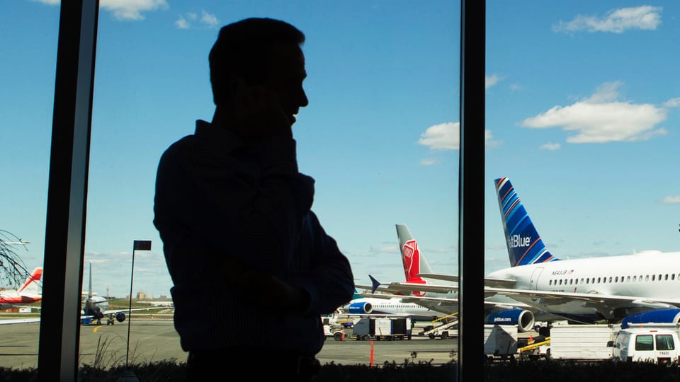 Person im Gegenlicht telefoniert vor einem Fenster auf einem Flughafen, im Hintergrund Flugzeuge