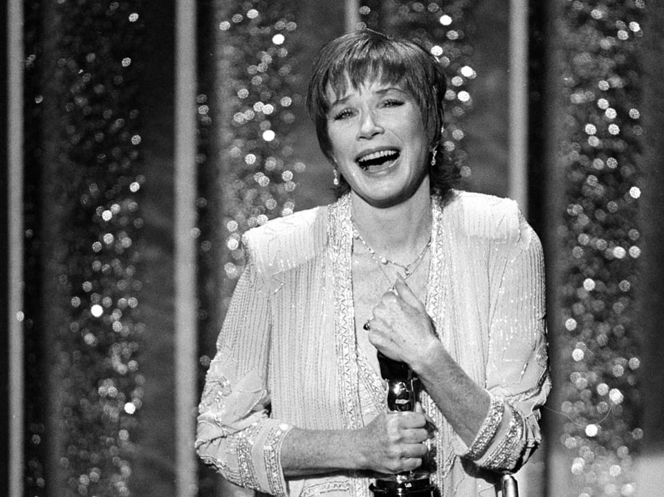 Schwarz-weiss Foto einer lachenden Frau mit Oscar auf einer Bühne mit Glitzerhintergrund.