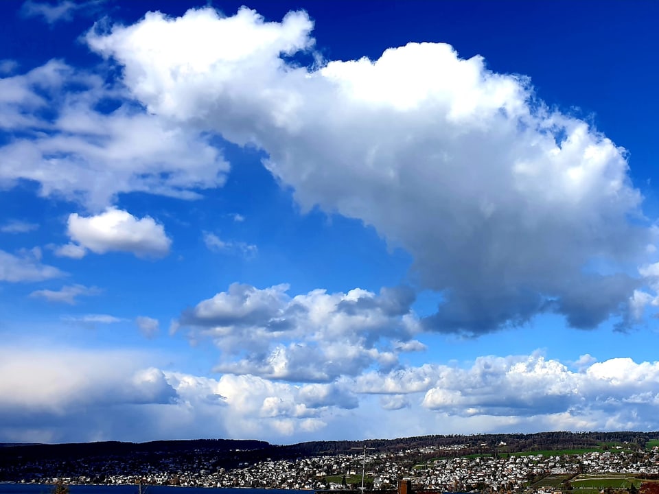 Am blauen Himmel sind grosse weisse Quellwolken vorhanden, darunter der Ort, umgeben von Hügeln.