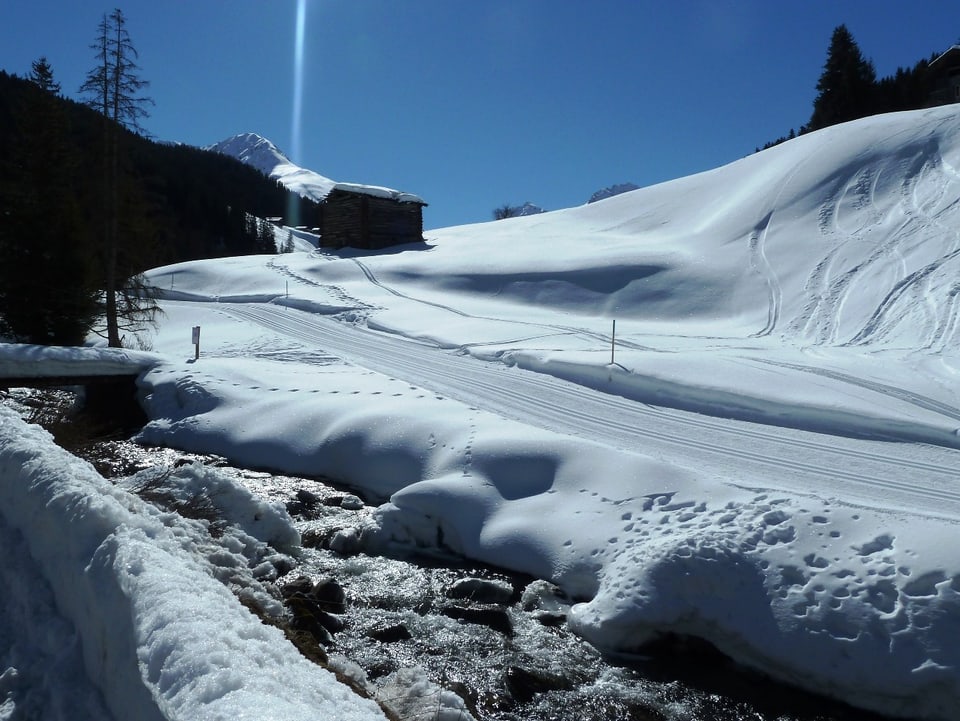Eine tiefverschneite Landschaft mit einem rauschenden Bach im Vordergrund. Die Loipe führt bergauf ins Sertigtal. Darüber stahlblauer Himmel.