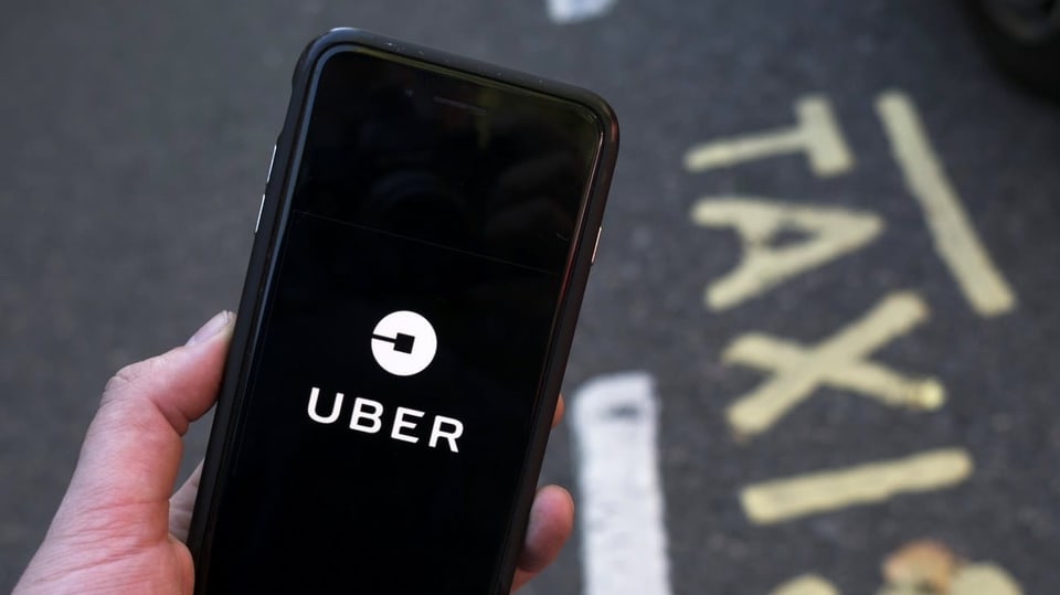 Eine Hand hält ein Handy, auf dem Display erscheint die App von Uber. Am Boden steht in gelber Schrift "Taxi". 