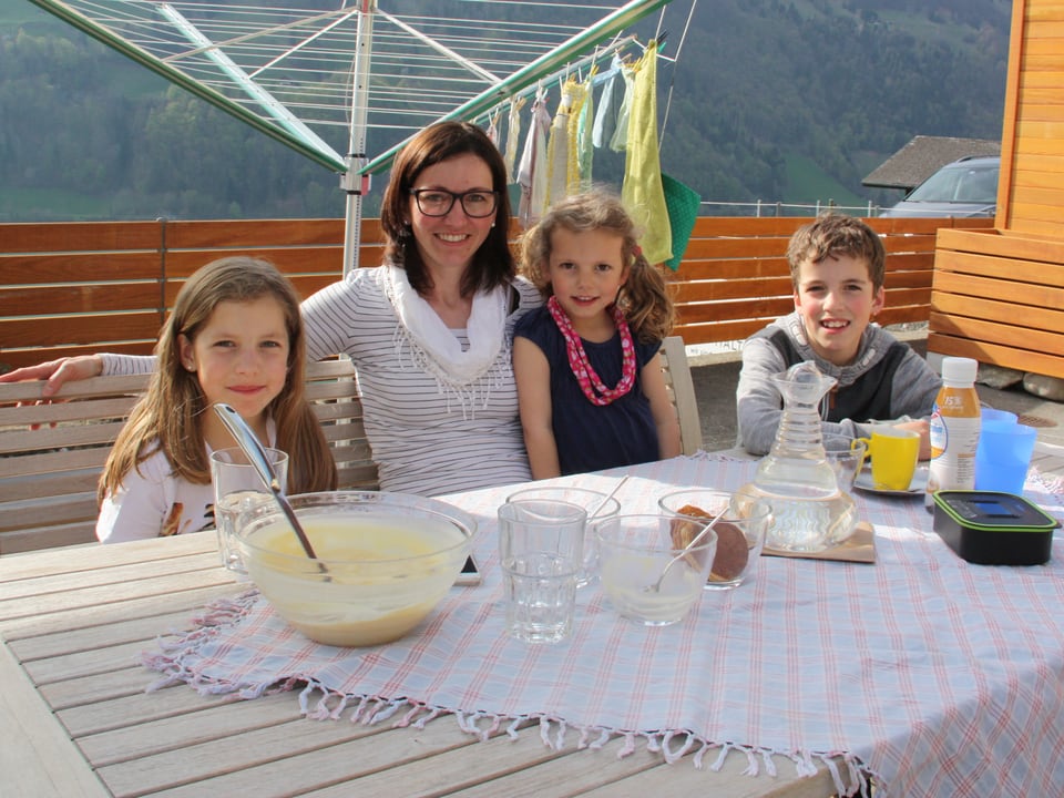 Eine Mutter sitzt mit ihren drei Kindern auf einer Terasse vor einem gedeckten Tisch.