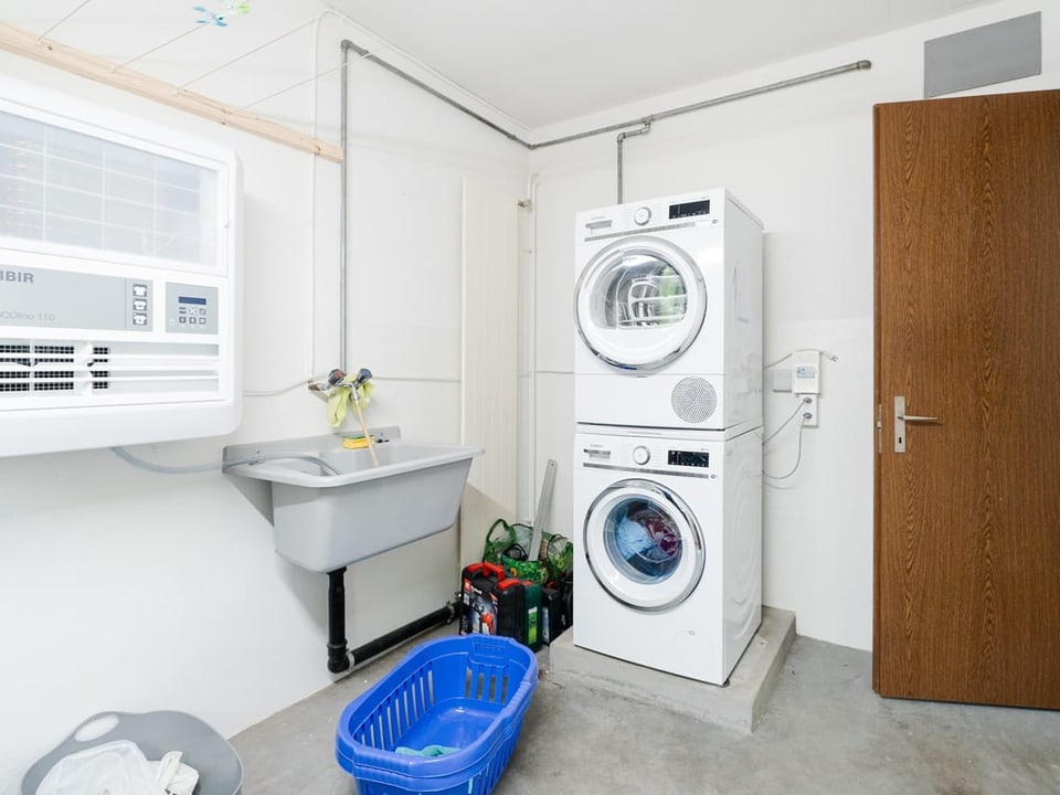 Ein Raum mit einem Waschturm. Dazu ein blauer Wäschekorb im Vordergrund und ein Lavabo an der linken Wandseite.