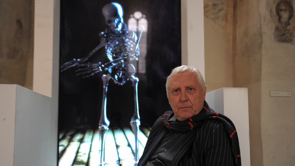 Peter Greenway sitzt vor einer seiner Projektionen, auf denen ein tanzendes Skelett erscheint.