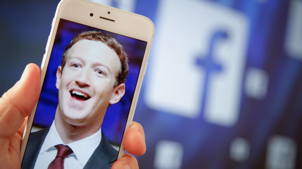 Symbolbild: Aufnahme Zuckerbergs auf einem Handy vor einem grossen Facebook-Logo.