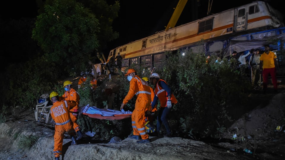 Rettungskräfte tragen Leichensack weg. Im Hintergrund das Zugswrack. Es ist dunkel.