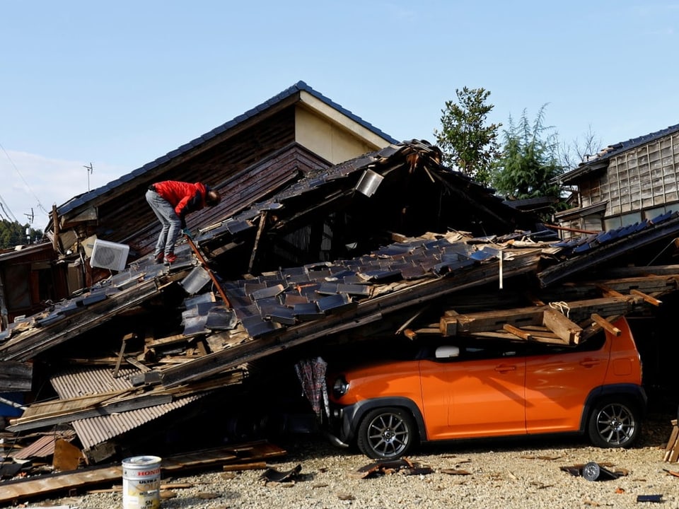 Mann steht auf dem Dach eines eingestürtzen Hauses. Unter den Trümmern steht ein Auto.