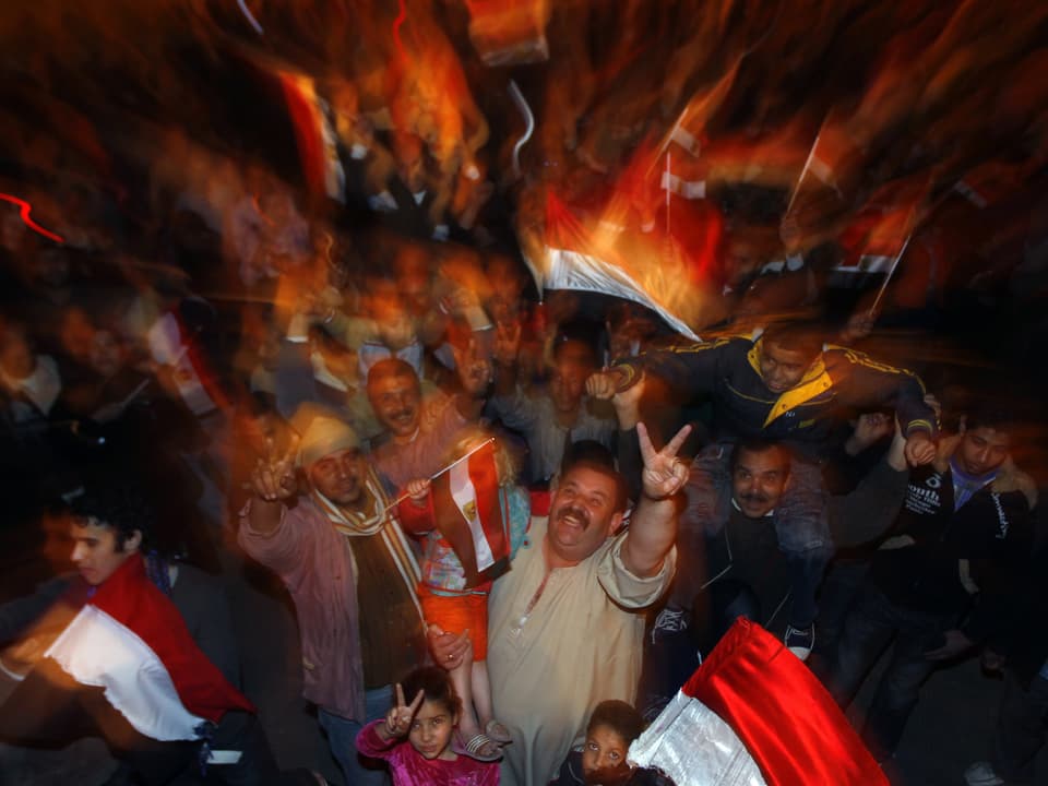 Jubelnde Menschen in der Nacht mit Flaggen. Mann in der Mitte macht das Victory-Zeichen.