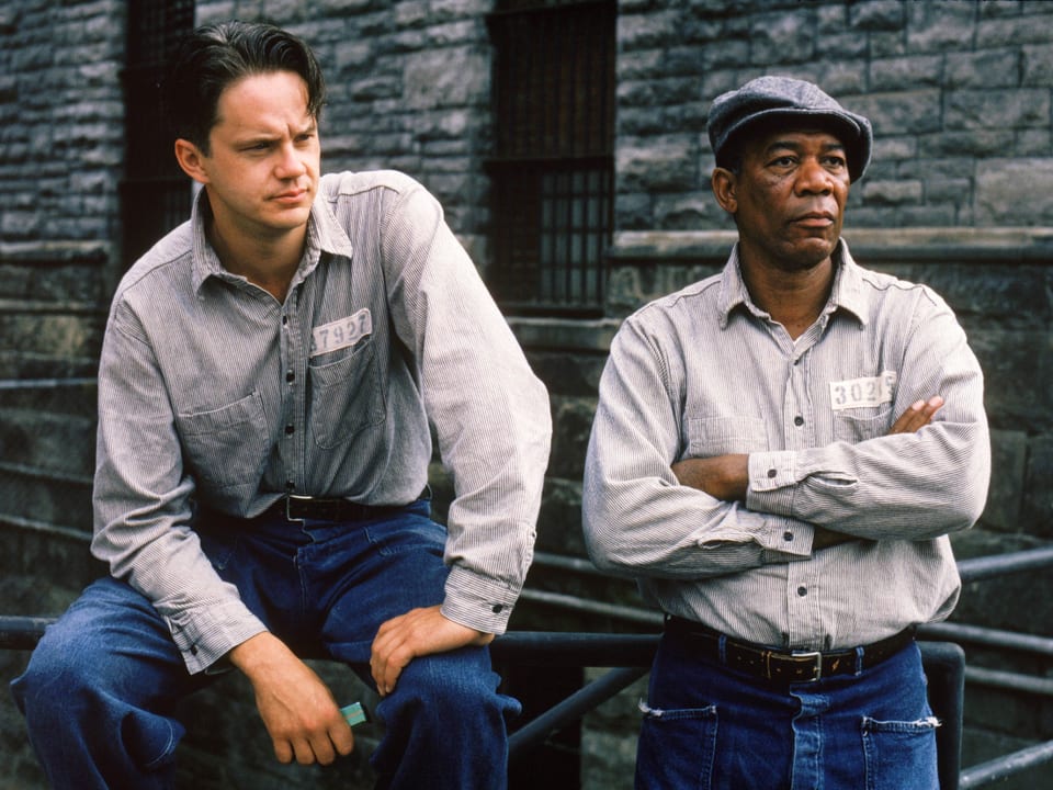 Zu sehen sind die Schauspieler Tim Robbins und Morgan Freeman in einem Gefängnishof. Sie tragen Sträflings-Kleidung. Ein Hemd mit einer Gefangenen-Nummer.