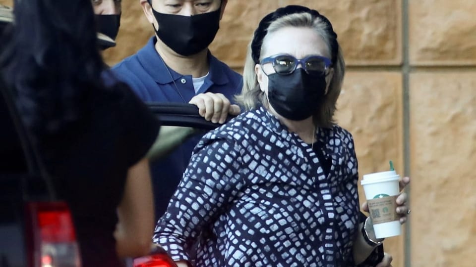 Hillary Clinton mit Sonnenbrille, Schutzmaske und Kaffeebecher in die Klinik gehend.