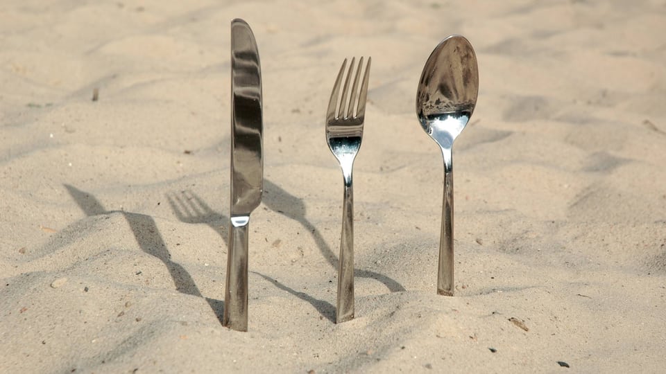 Messer, Gabel und Löffel stecken in Sand.