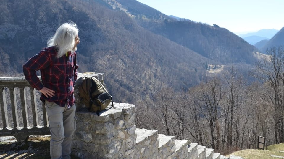 Links Mann mit langen, grauen Haaren schaut über die Hügel, steht oben an Treppe.