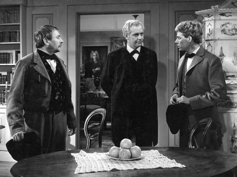Szene in einer Stube. Drei Männer stehen im Raum und sprechen miteinander. Vor ihnen steht ein Tisch, auf dem Tisch in einer Früchteschale liegen fünf Äpfel.