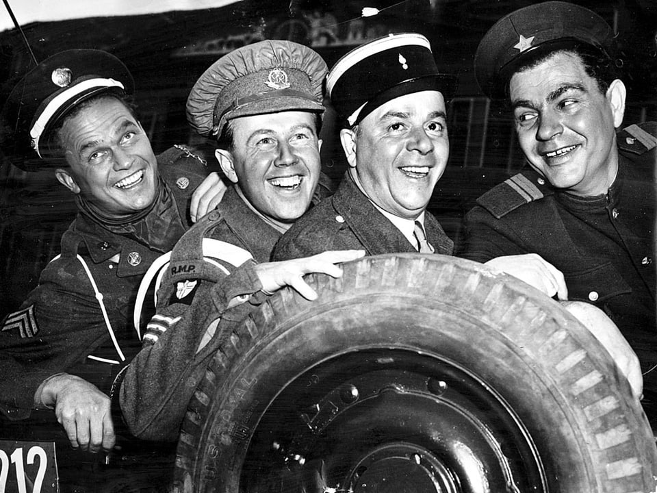 Vier uniformierte Männer stehen lachend hinter einem Pneu.