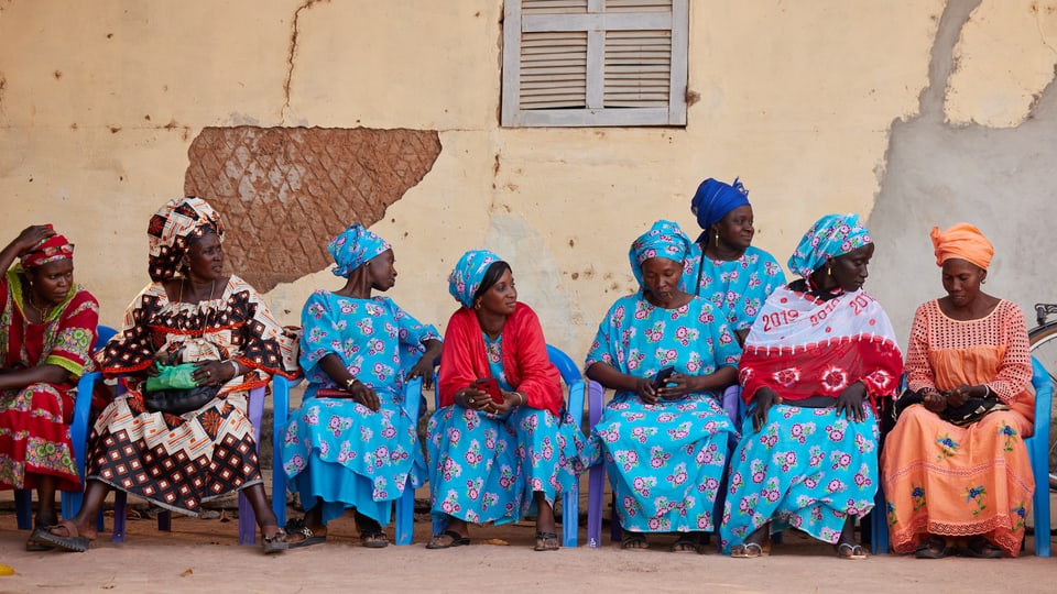 Frauen im Senegal in farbigen Kleidern sitzen auf Stühlen vor einer Wand.