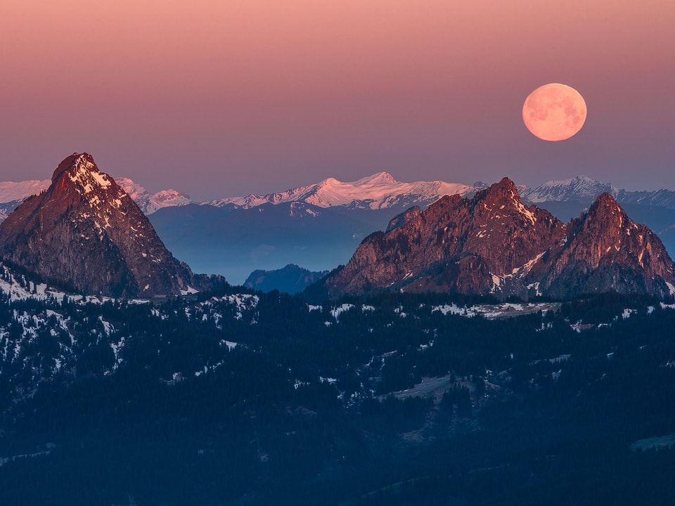 Der beinahe volle Mond geht hinter den Bergen unter. Im Morgenlicht erhält der Himmel eine rötliche Farbe.