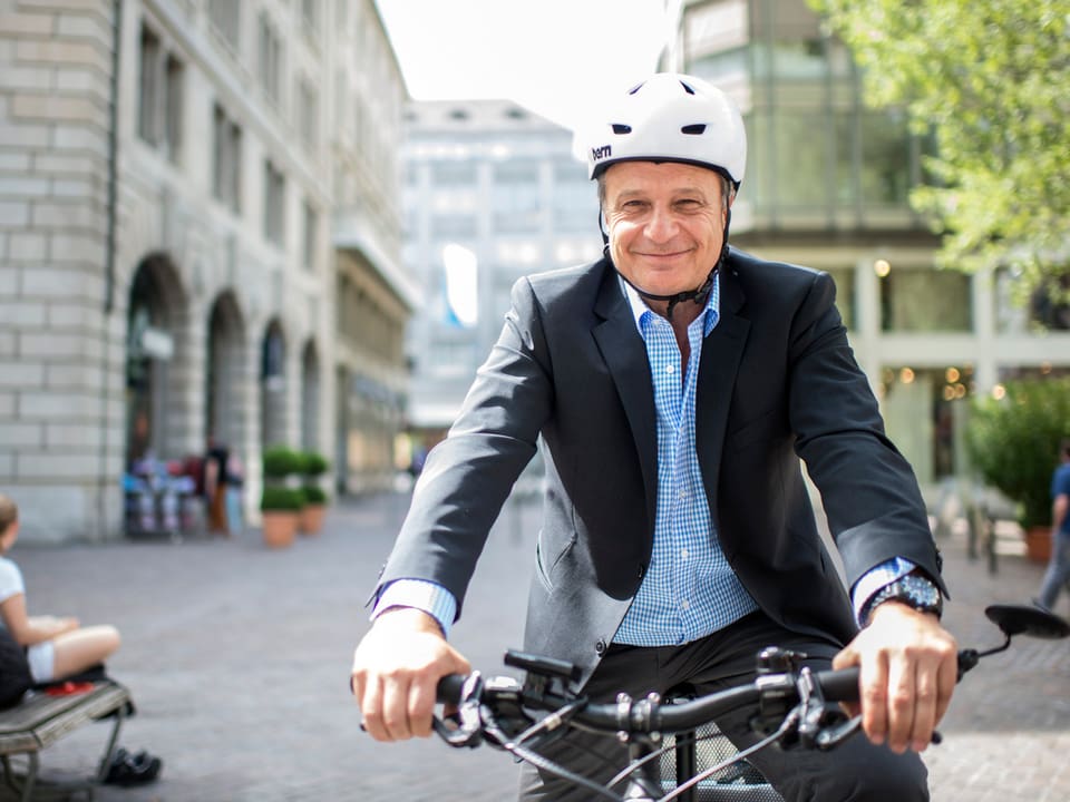 Ein Mann sitzt auf einem Fahrrad, mit weissem Helm und im Anzug. Beide Hände fest am Lenker und ein Lächeln in die Kamera.