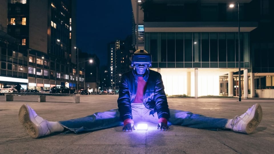 A man wearing virtual reality glasses and a mobile phone sitting on the ground in a city at night.