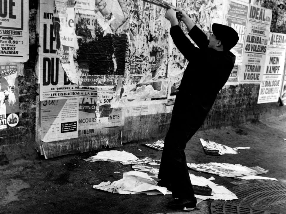Eine schwarz-weisse Fotografie zeigt einen dunkel gekleideten Mann, der Plakatwände abreisst.