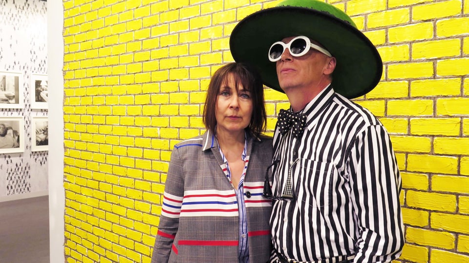 Ein Mann und eine Frau vor einer gelben Wand. Der Mann trägt einen grossen Hut und eine grosse Sonnenbrille. 