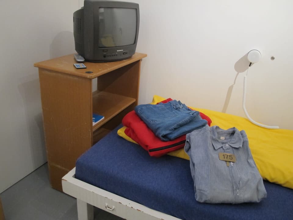 Eine Gefängniszelle mit TV, Bett, Kleidung auf dem Bett.