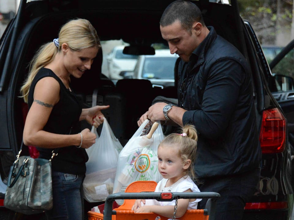 Michelle Hunziker (links im Bild) und ihr Bodyguard (rechts im Bild) stehen vor dem Kofferraum eines schwarzen Autos und räumen die Tüten ein. Sole wartet im Einkaufswagen (im Vorderdrund).