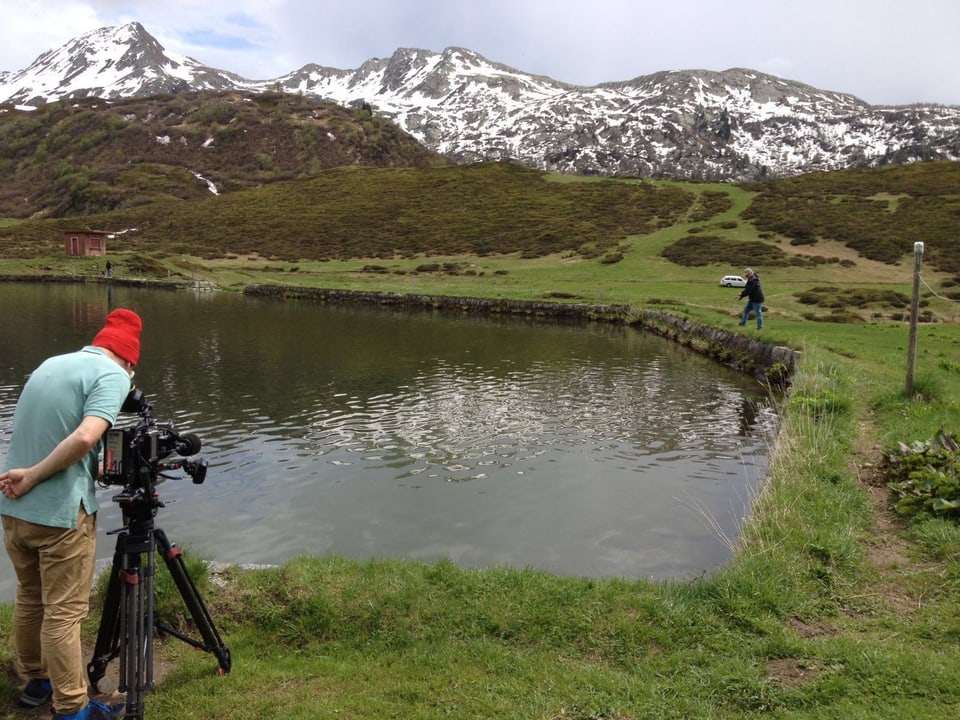Kameramann mit roter Mütze filmt am Ufer eines Beergsees, auf der anderen Seite des Sees steht ein Mann mit Fischerrute. 
