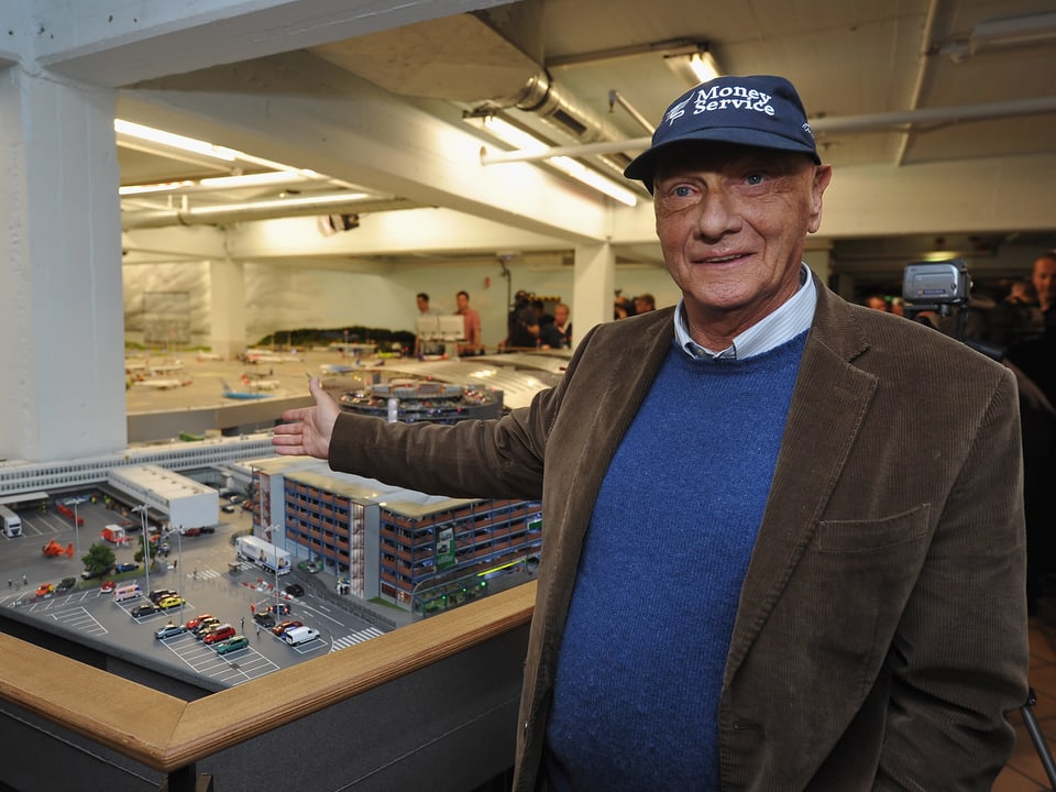 Niki Lauda mit blauer Kappe zeigt auf einen Modellplan eines Hauses