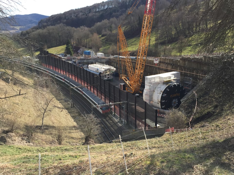 Die bisherige Zugsstrecke mit einem Zug der in den alten Tunnel fährt, daneben steht die Tunnelbohrmaschine.