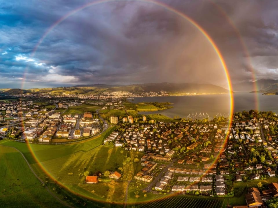 Luftbild mit einem runden Regenbogen über Dorf am See mit dunkeln Wolken am Himmel. 