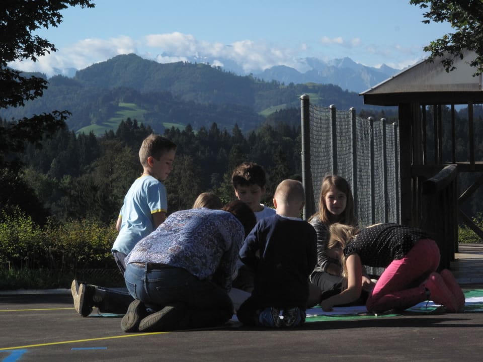 Kinder knien auf dem Pausenplatz um ein grosses Blatt Papier, im Hintergrund Wälder, Hügel und Berge.