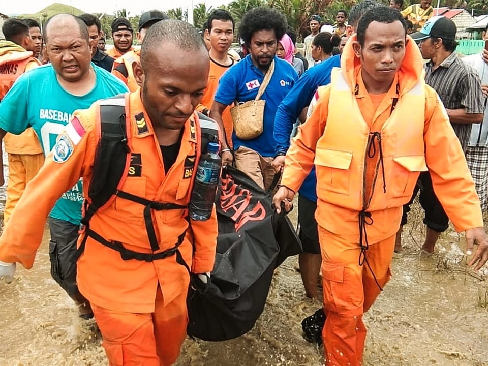 Rettungskräfte in orangen Anzügen tragen ein Opfer in einer Tasche. 