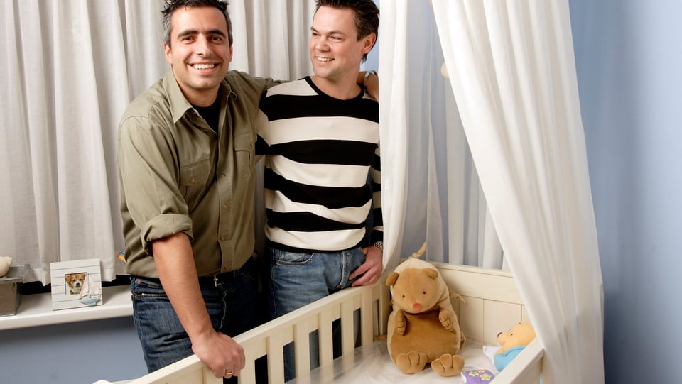 Eing gleichgeschlechtliches Männerpaar steht neben einem Kinderbettchen.