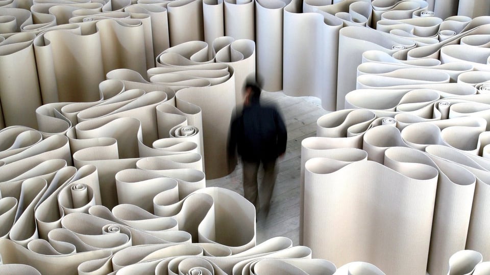 Mann geht durch ein Labyrinth aus hochkant aufgestellten Papierbögen.