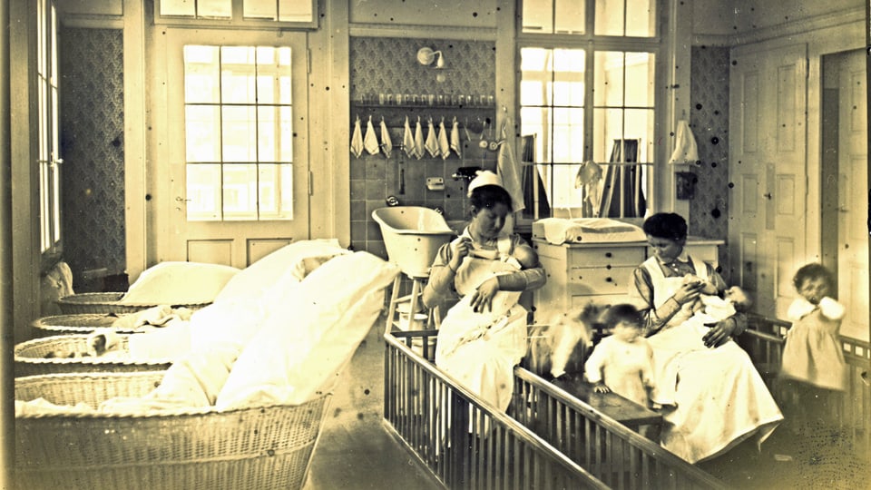 Aufnahme von ca. 1930, wo zwei Kinderpflegerinnen mit weissen Schürzen und Häubchen zwei Babys den Schoppen geben, während etwas grössere Kinder um sie herum springen. 