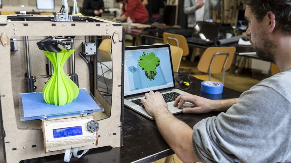 Ein Mann arbeitet am Computer in einem Labor. Neben ihm steht ein 3D-Drucker mit einer grünen Figur.