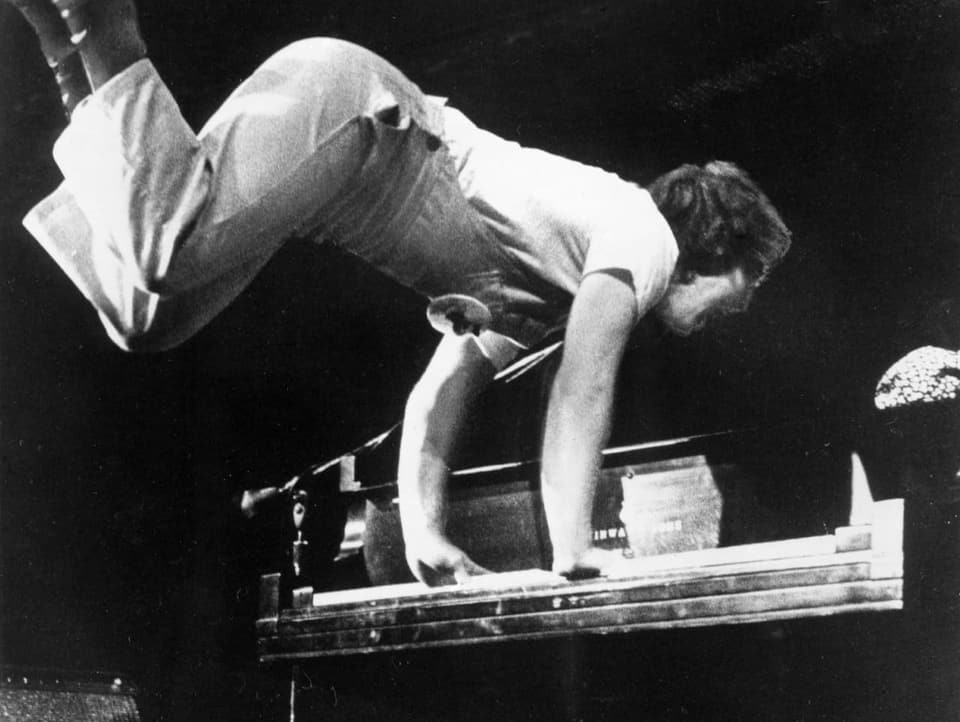Fotoaufnahme eines jungen Mannes, der sich mit beiden Händen auf dem Klavier abstützt und seine Beine in die Höhe wirft.