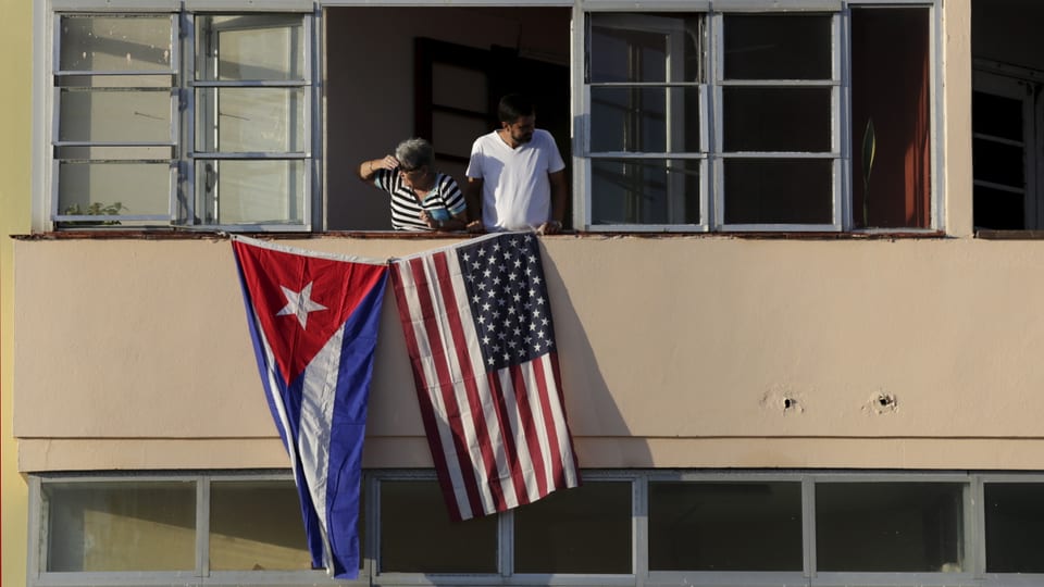 Eine kubanische und eine amerikanische Flagge an einem Haus, aus dem Fenster schauen zwei Personen.