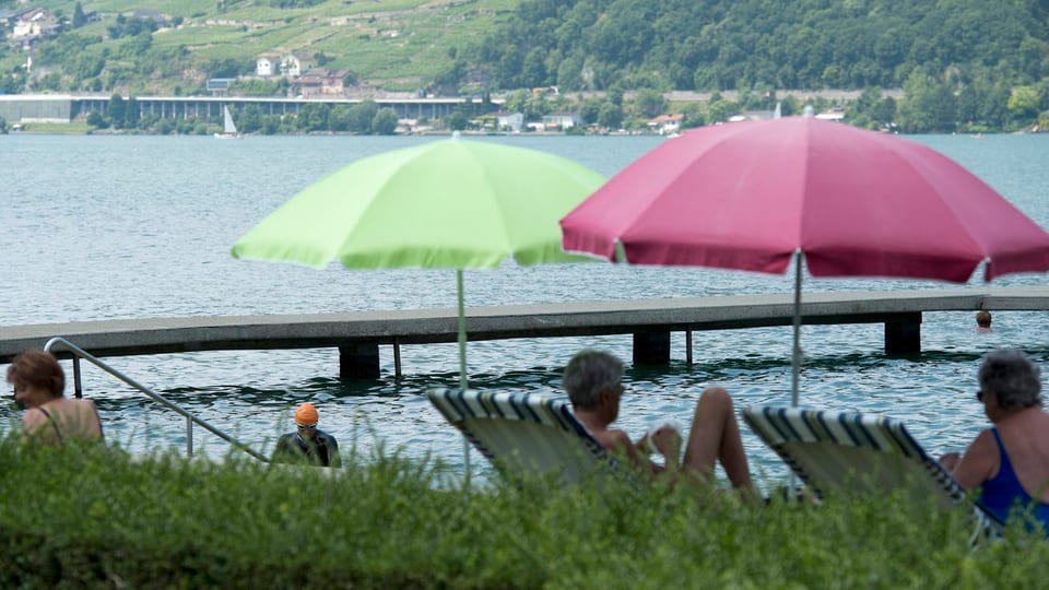 Bielerseeufer mit Steg, zwei besetzte Liegestühle mit Sonnenschirmen.