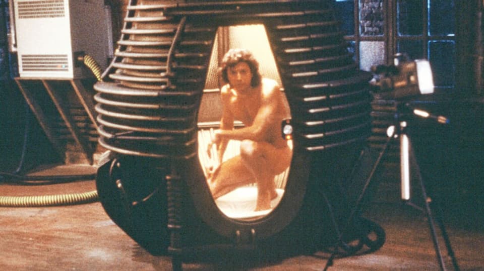 Ein nackter Mann kniet in einer futuristisch anmutenden Kapsel.