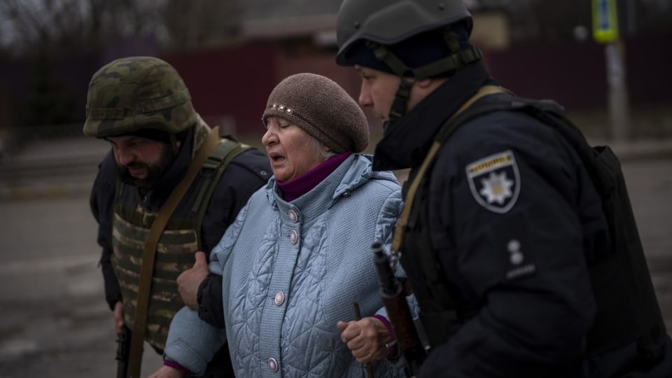 Ukrainische Polizisten helfen einer Frau auf der Flucht aus Irpin. Im Hintergrund sind Raketen zu hören.