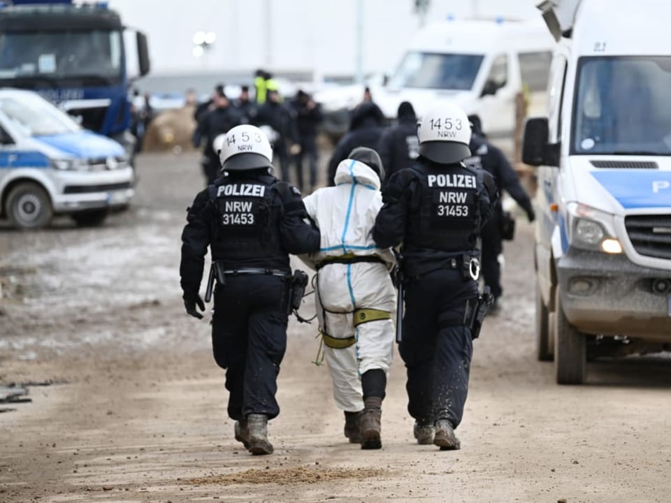 Eine Person steht zwischen zwei Polizisten. Die Polizisten haben die Person an den Armen gegeriffen.