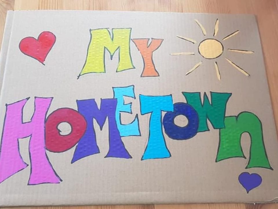 «My Hometown» Plakat auf Karton auf Holzboden. Schriftzug ist farbig und oben rechts ist eine Sonne aufgemalt.