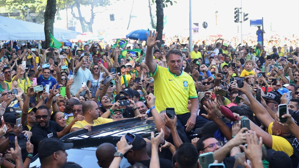 Jair Bolsonaro trägt ein gelbes Brasilien-Shirt und sitzt auf dem Dach eines Autos, umringt von zahlreichen Anhängern.