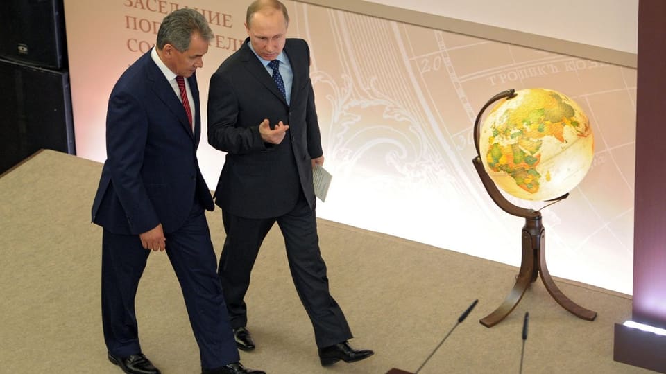 Putin und sein Verteidigungsminister gehen Seite an Seite an einem von innen beleuchteten Globus vorbei.