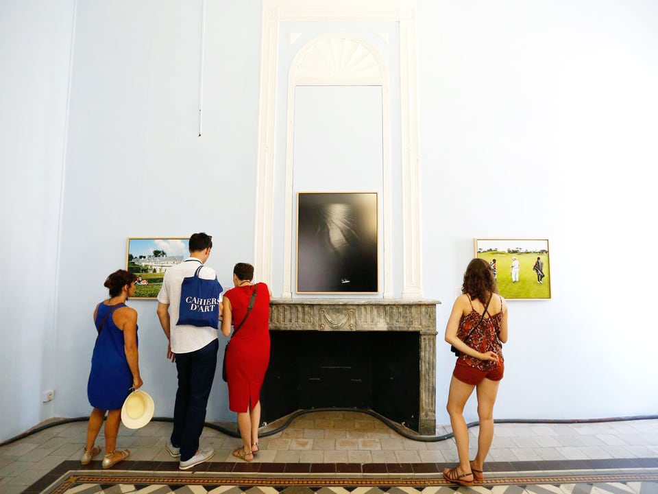 Besucher betrachten zwei Fotografien in einem Raum, der einen reich verzierten Steinboden hat. An der Wand ein Kamin. 