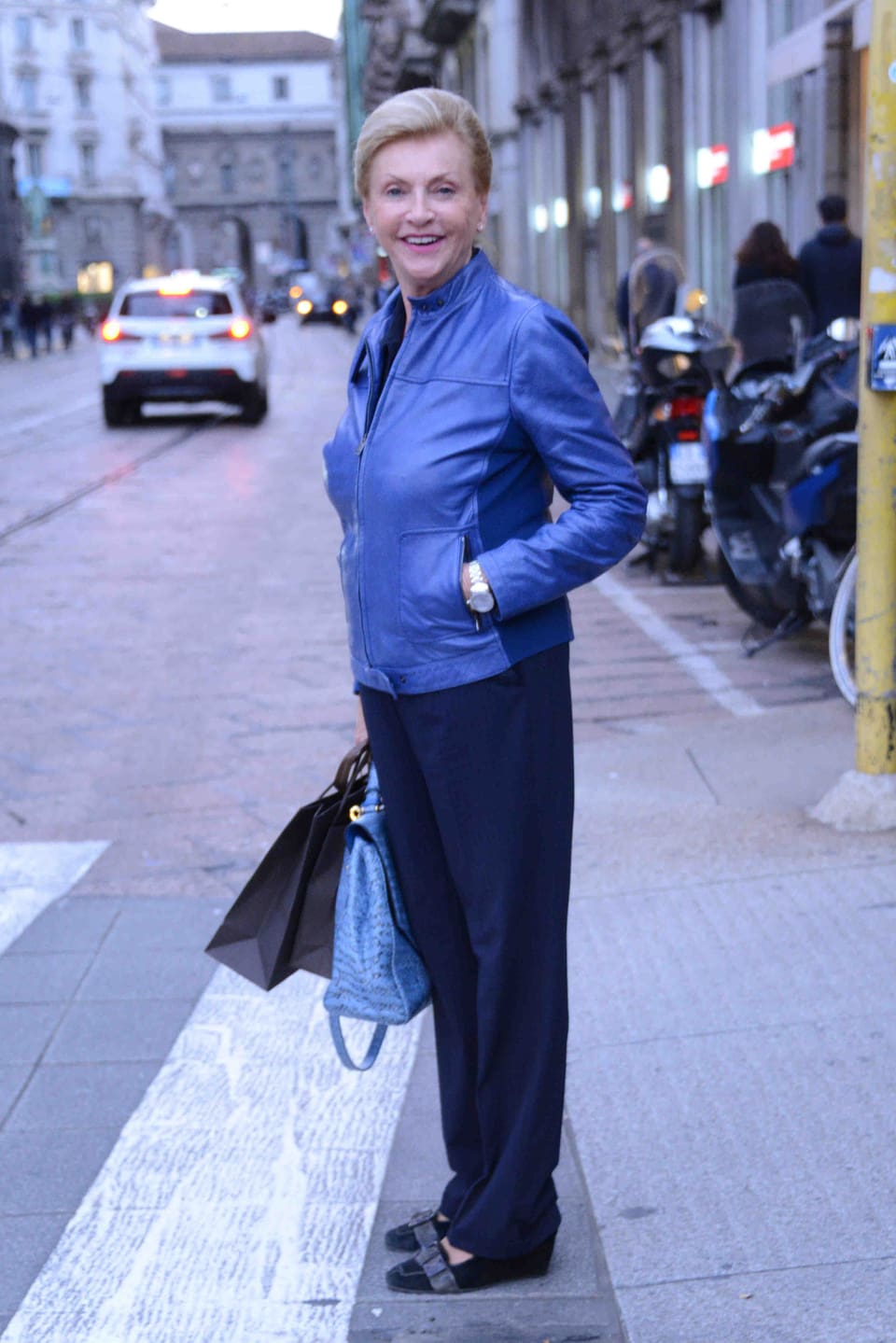 Michelle Hunzikers Mutter in einer blauen Lederjacke vor einem Fussgängerstreifen stehend und die Kamera lachend. Sie trägt zwei Taschen.