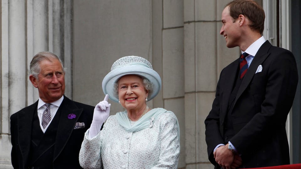 Queen auf einem Balkon, sie winkt. Daneben Charles und William.