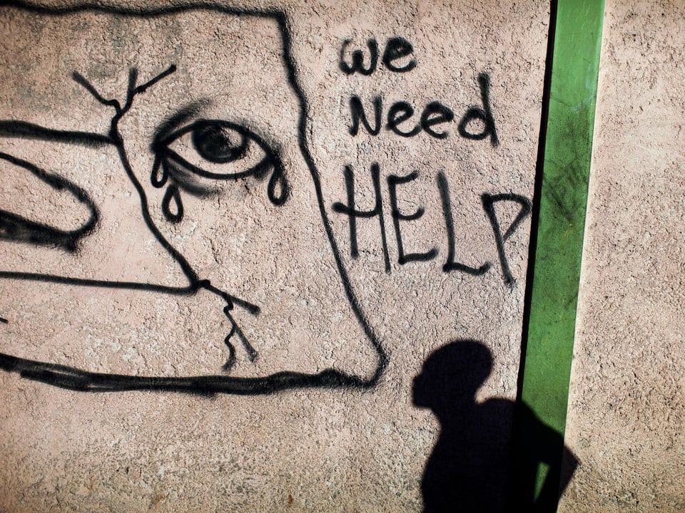 Silhouette eines Mannes, der vor einer Hausmauer steht. Auf der Mauer ein Grafiti mit der Aufschrift «We need help».