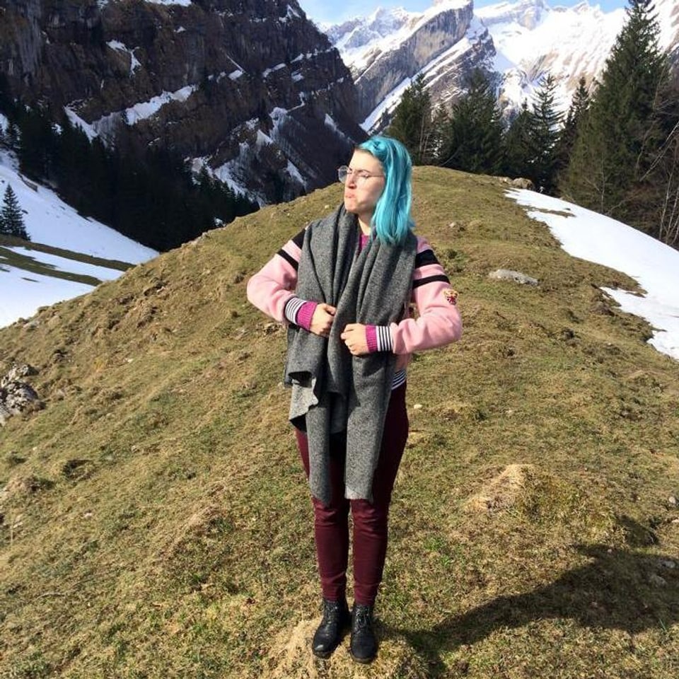 Eine junge Frau mit blaugefärbten Haaren steht auf einem Berg und macht eine Pose wie ein Muskelprotz. 